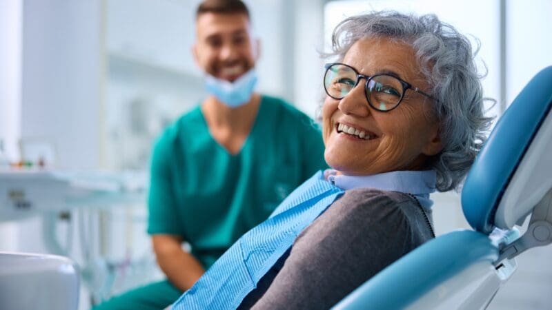 Happy senior woman at dental clinic looking at camera. Her dentist is in the background.